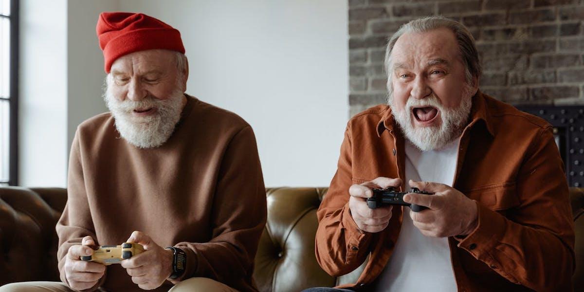 Två glada pensionärer sitter i en soffa och spelar tv-spel.