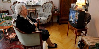 En äldre kvinna tittar på tv med fjärrkontrollen i handen. Många äldre tittar på på tv-program när de sänds, ofta på dagtid och tidig kväll och en krönikör tycker att SVT borde tänka mer på dem