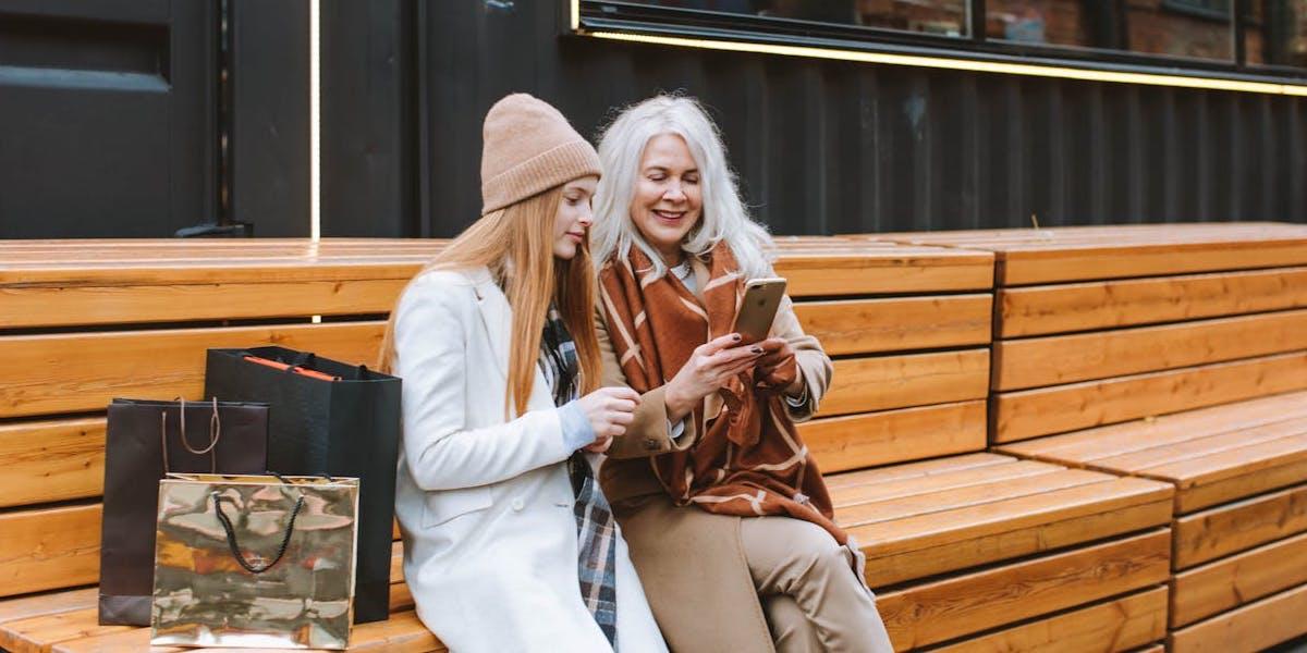 En yngre och en äldre kvinna sitter på en bänk och tittar i en mobiltelefon.