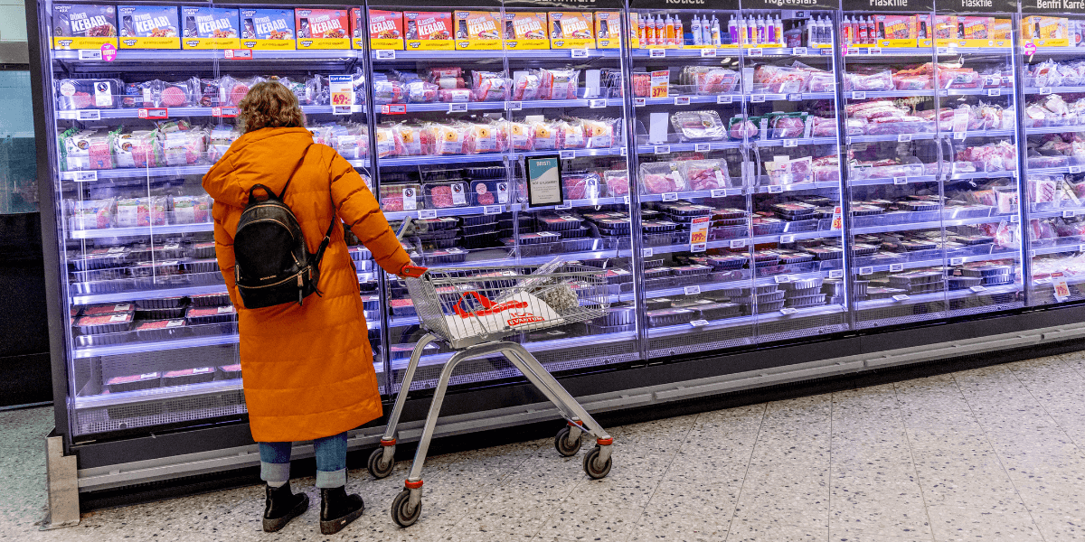 En kvinna i orange vinterjacka som undrar över matmomsen står framför en kyl med köttvaror i en matbutik.
